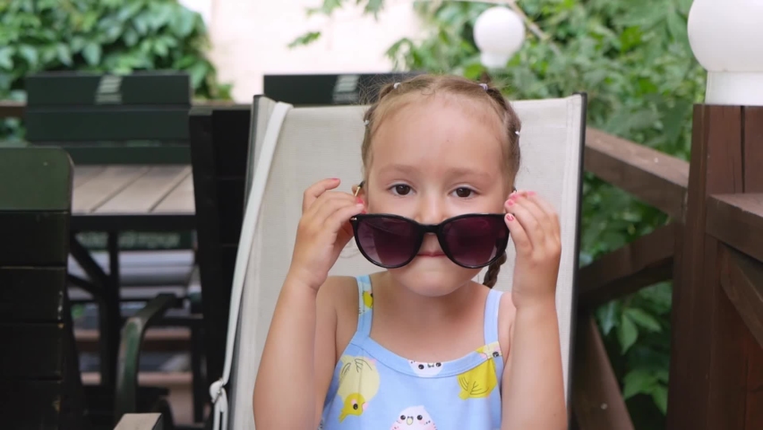Portrait of happy smiling toddler girl in sunglasses in summer café. Little girl has fun and fools around with woman sunglasses in sunny day.