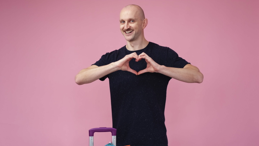 Holiday joy. Inspired man. Send love. Happy funny attractive guy with suitcase showing heart gesture on chest on pink background.