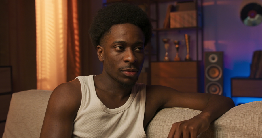 Golden hour sunshine man is sitting on sofa in living room smiling contentedly looks ahead laughing, good mood, guy is watching TV, his favorite series, relaxing in home clothes.