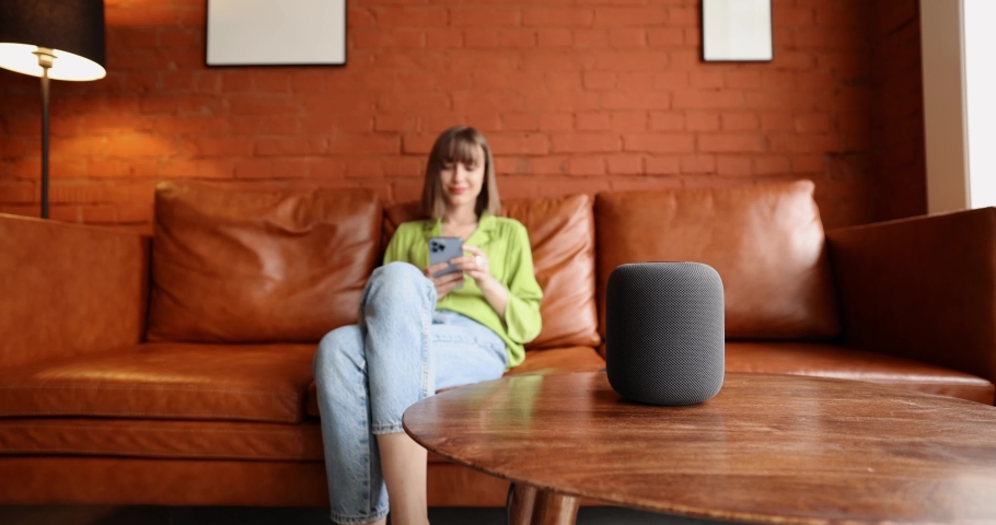 Young woman using phone and controlling home devices with a voice command while sitting relaxed on leather couch. Concept of smart home and leisure time with gadgets Royalty-Free Stock Footage #1092421617