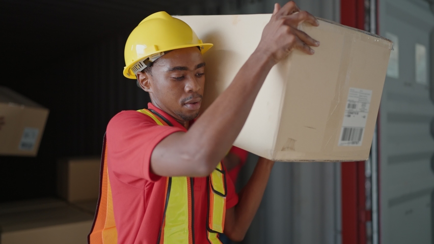 Young African American black workers courier lifting cargo boxes stacking on pallet in warehouse wearing engineer suit and helmet for safety. Shipment boxes. Warehousing storage.