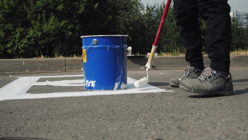 Close-up of manual utility male worker dipping roller brush in paint metal bucket, painting white marking line of disabled car parking lot sign on asphalt, drawing on road surface, summer daytime.