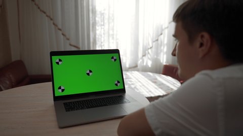 Man watches movie on laptop sitting at wooden table in room against bright window. Modern computer with green chromakey screen close view