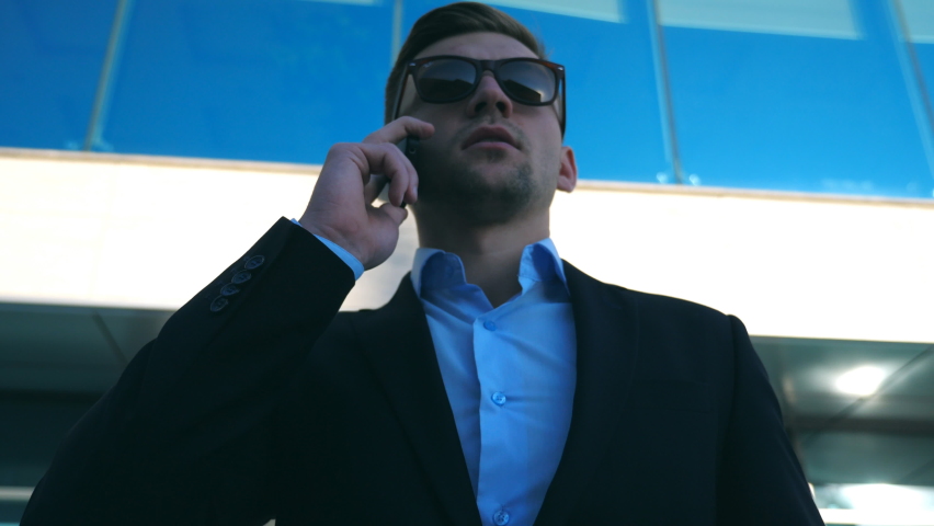Portrait of young businessman in sunglasses talking on phone outdoor near office. Close up of handsome business man speaking on cellphone with modern business building at background. Slow motion