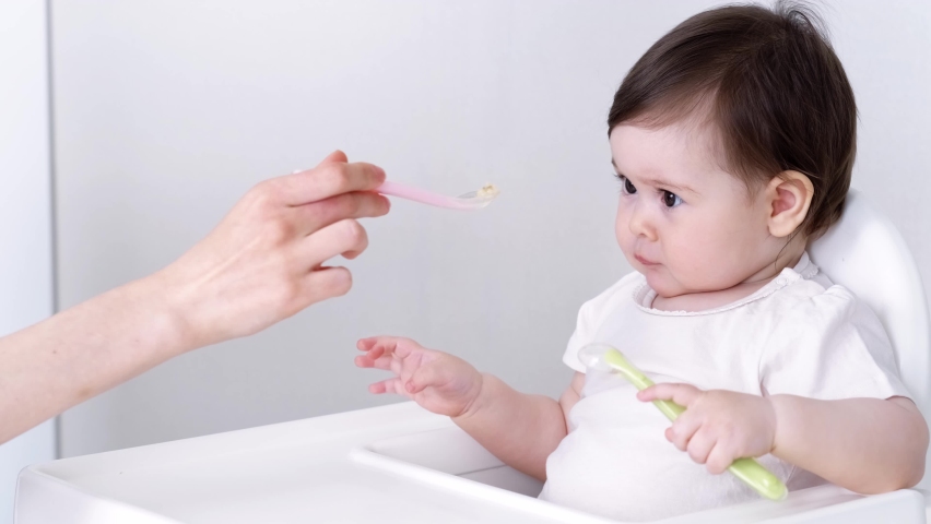 Baby girl eating blend mashed food sitting, on high chair, mother feeding child, hand with spoon for vegetable lunch, baby weaning, first solid food for young kid. Royalty-Free Stock Footage #1092435991
