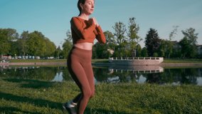 Young sportive female doing reverse lunges at summer nature park. Female person wearing orange sport clothes exercising outdoors, doing squats with the morning sunlight. Bodyweight training exercise