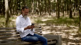 An adult man with a gray beard on a park bench works with a phone. 4K slow motion video.