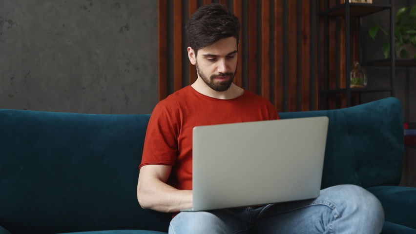Young happy minded man wears red t-shirt hold type use work on laptop pc computer sits on blue sofa couch stay at home flat rest spend free time in living room indoors grey wall. People lounge concept