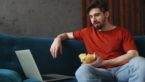 Young smiling man wears red t-shirt watch movie film on laptop pc computer eat chips sits on blue sofa stay at home rest relax spend free time in living room indoors grey wall. People lounge concept