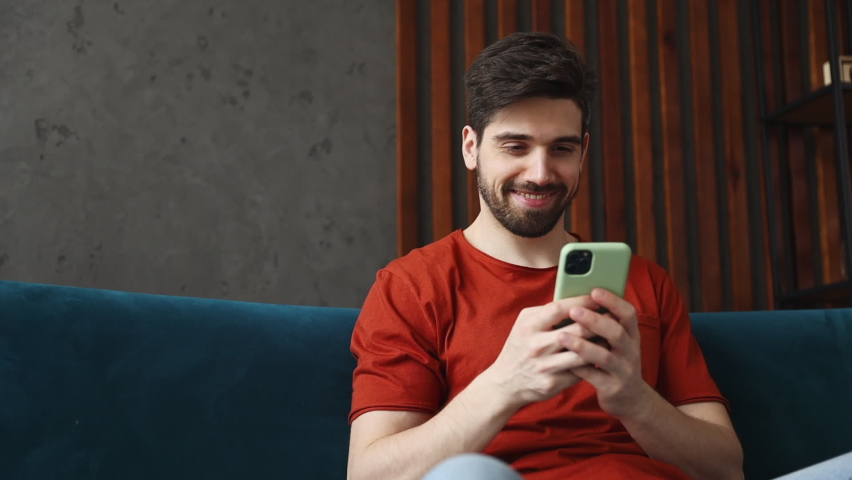 Young man wears red t-shirt hold mobile cell phone watch internet memes sit on blue sofa couch stay at home hotel flat rest relax spend free time in living room indoors grey wall People lounge concept Royalty-Free Stock Footage #1092442361