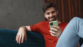Young man wears red t-shirt hold mobile cell phone watch internet memes sit on blue sofa couch stay at home hotel flat rest relax spend free time in living room indoors grey wall People lounge concept - Powered by Shutterstock - Get 15% off with code: PIKWIZARD15