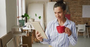Video of happy biracial female designer using tablet and drinking coffee in studio. Art, crafts, creativity, creation process and running own business concept.