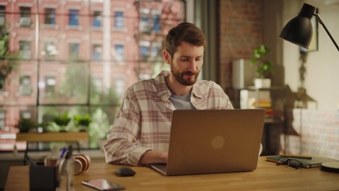 Stylish Bearded Young Adult Man Using Laptop Computer in Creative Loft Office Space. Happy Specialist in Checkered Shirt Working Remotely on Agency Projects from Home. Arc Shot.