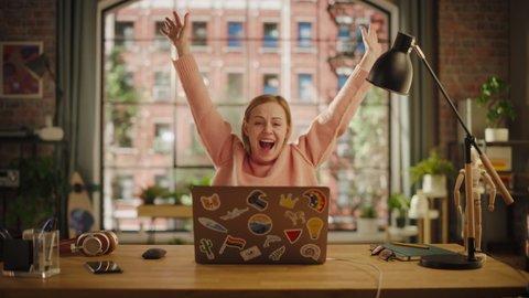 Happy Young Woman Working on Laptop Computer while Sitting in Stylish Creative Office. Successful Freelancer Receiving New Order and Celebrating. Female Winning Lottery and Raising Hands with Joy.