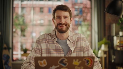 Portrait of a Handsome Bearded Young Adult Man in Casual Clothes Using Laptop Computer in Stylish Loft Apartment at Home. Happy Specialist in Checkered Shirt Looking at Camera, Smiling.