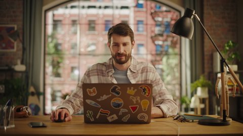 Handsome Bearded Man Working on Laptop Computer while Sitting at a Table in Stylish Cozy Loft Living Room. Freelancer Working From Home. Browsing Internet, Using Social Networks, Being Productive.