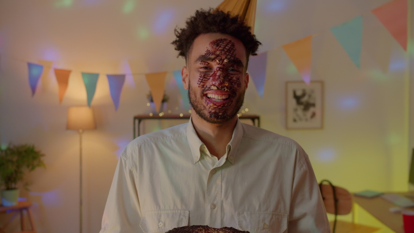 Slow motion portrait of funny young man with chololate covered face eating birthday cake at party in decorated apartment. Youth and celebrations concept.