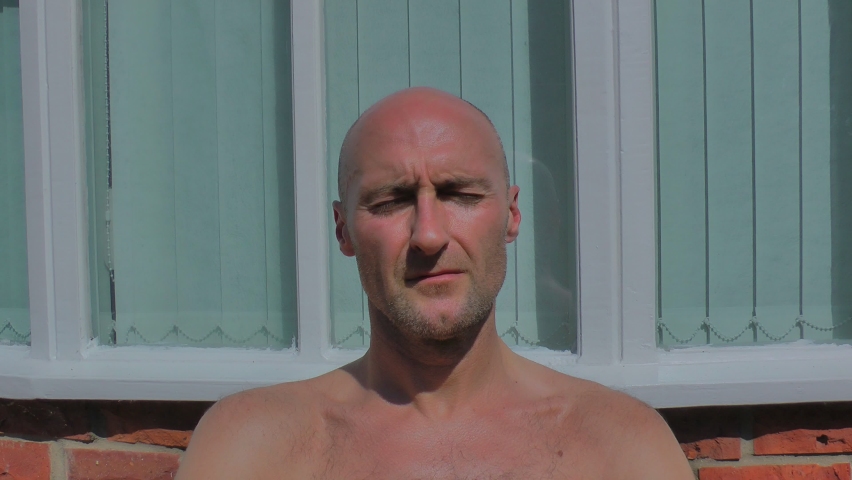 A middle aged bald man sits outside in the uncomfortably hot summer sun. Sun burnt and sweating, he wipes his brow of sweat. Filmed in slow motion. Royalty-Free Stock Footage #1092464241