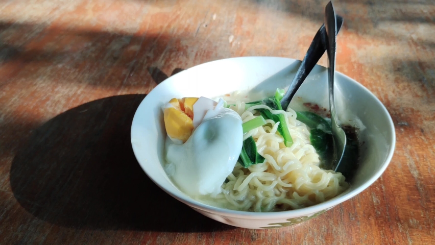 Top view of instant noodle with egg in white bowl, popular food in Indonesia. Served on wooden motif table. Negative space Royalty-Free Stock Footage #1092466531