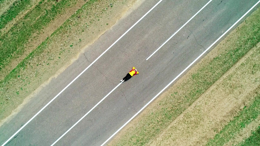 Aerial top down view of a young woman with pink hair braids and yellow jacket laying on an asphalt road.
