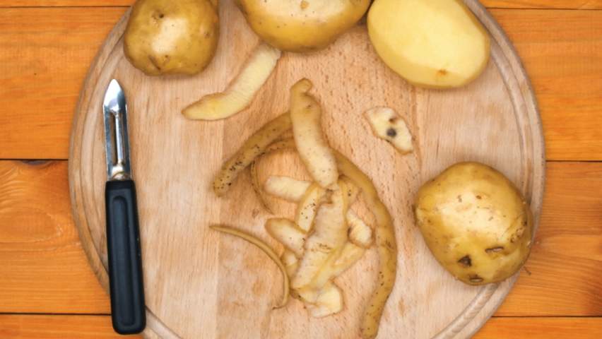 The cook is peeling potatoes. A man peels raw fresh potatoes with a manual potato peeler. Peeled potatoes and peel on a cutting board on a wooden table. Cooking food in the kitchen. Close-up. Top view Royalty-Free Stock Footage #1092466875