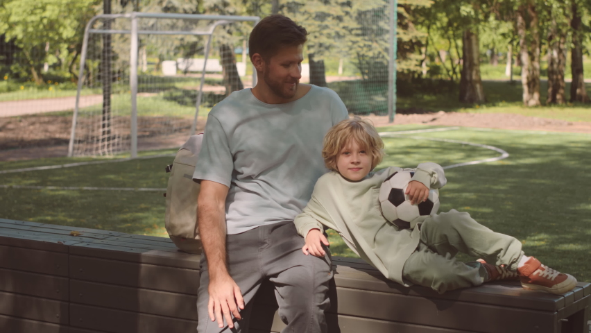 Slowmo portrait of Caucasian father and his 7 year old son resting on bench after football training on field in park, smiling at camera
