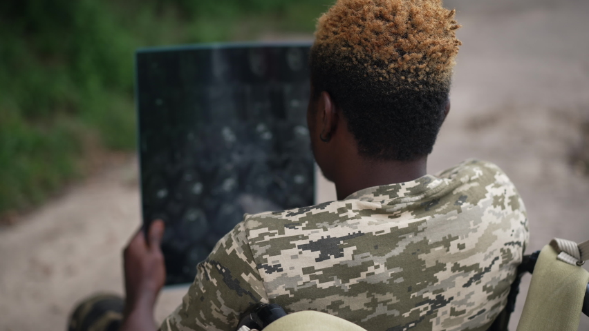 Shooting over shoulder of young man in wheelchair with X-ray image thinking sitting outdoors. African American sick soldier analyzing medical result in summer park forest. Slow motion