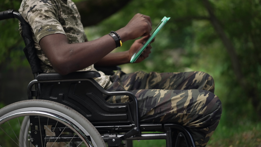 Side view unrecognizable African American man in wheelchair messaging online on tablet outdoors in slow motion. Young brave soldier surfing Internet resting in green summer park