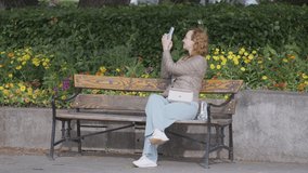 A picturesque park. Action. A young woman with curly hair with her legs crossed takes pictures of landscapes on her phone while sitting on a bench.