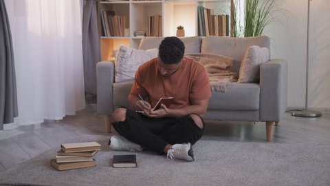 Home study. Learning hobby. Creative education. Smart guy sitting cross-legged with books on floor taking notes at modern living room interior.