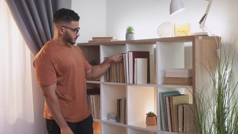 Home library. Reading hobby. Learning lifestyle. Curious smart guy in glasses checking book collection on bookshelf at modern living room interior.