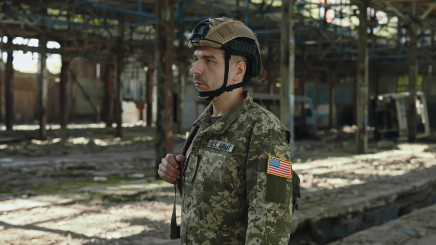 American servicemen turning head and looking at camera while standing with black backpack on abandoned factory. Man wearing protective helmet and camouflage military uniform. Royalty-Free Stock Footage #1092478159
