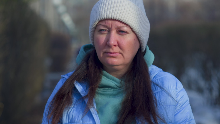 Portrait Of Serious Brunette Woman Is Looking Down Walking Outdoors On The Street. She Is Wearing Knitted Hat, Blue Jacket And Hoodie Early Spring. Tired Person Thinking Something Worried.