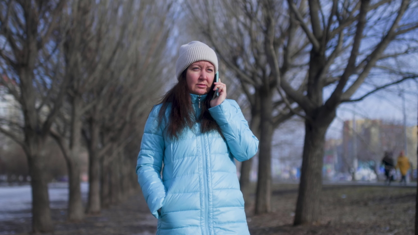 Woman Is Talking On Mobile Phone In Early Spring Park Near The Naked Trees. She Is Wearing Hat And Jacket. Brunette Woman Is Calling On Her Smartphone During Her Walk In City Park. 