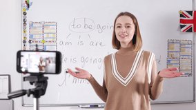 Female english teacher leading video lesson online while standing in classroom. Caucasian woman in casual attire using modern smartphone with tripod for work.
