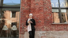 Attractive smiling muslim woman in hijab texting on smart phone standing on background of building wall from red brick. Portrait of young arabian businesswoman using mobile phone in city.