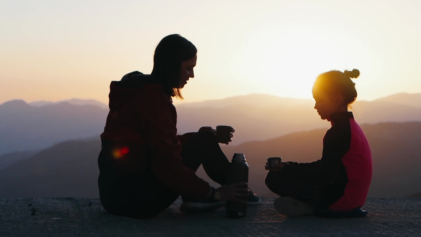 Young mother and 9 years daughter are spending time together at sunset on observation deck. They drinks tea with beautiful mountains view. Silhouette 4k footage.