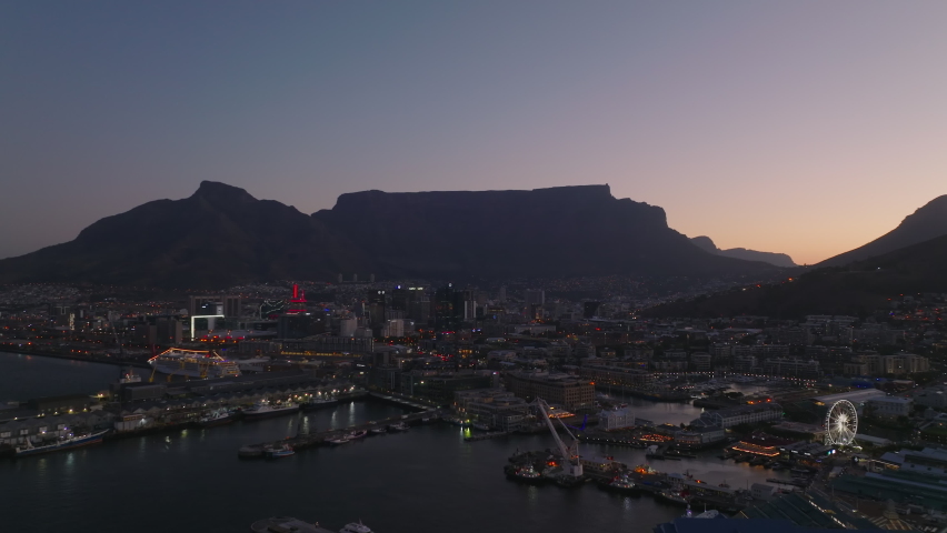 Fly above city marina at sea coast. Buildings and streets of evening town and silhouette of mountains against sky at dusk. Cape Town, South Africa