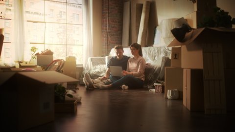 Moving Young Homeowners: Couple Sitting on the Floor of their New Apartment Use Laptop Computer for Online Shopping. Mortgage Loan, ecommerce, Planning Decoration of Sweet Home. Warm Nostalgia Colors