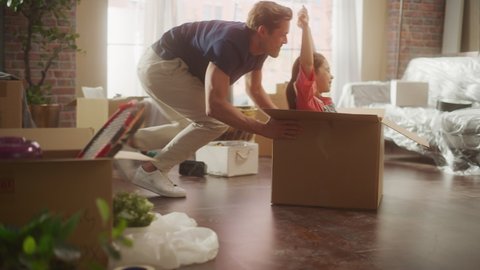 Moving in: Young Family of New Homeowners Has Fun. Father Driving Lovely Little Daughter in Cardboard Box. Dad and Pretty Cheerful Child Racing Together Through the Sunny Apartment Living Room.