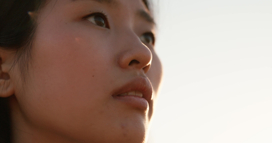 Close-up face of Asian woman, with brown eyes looking up outdoors. Beautiful young girl with black hair feeling the pleasure of rural landscape, standing in the background of a cloudless sky | Shutterstock HD Video #1092510259