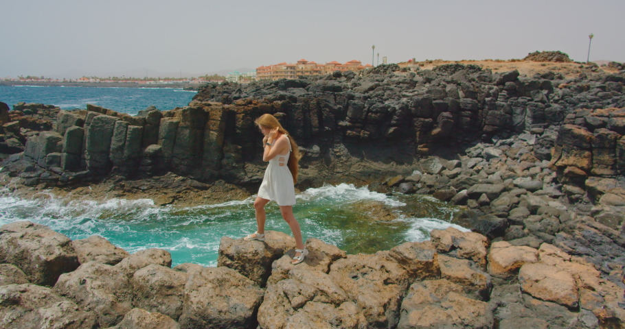 Side view of caucasian woman standing on rock in sea enjoying relaxing sunny day at the backview cityscape. Nature travel ocean big waves, outdoor rocky volcanic landscape, wind in slow motion.
