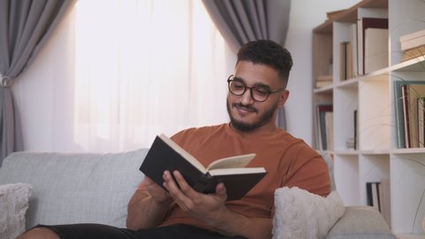 Reading hobby. Book lover. Weekend leisure. Relaxed curious smart guy in glasses enjoying funny story turning pages on home couch at modern interior.