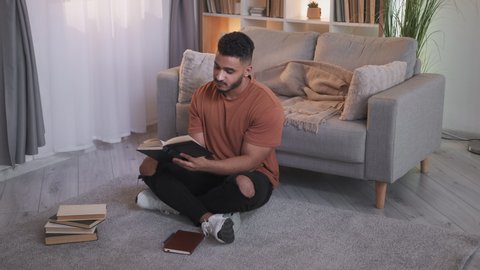 Book lover. Home reading. Weekend leisure. Curious intelligent guy sitting cross-legged on floor with literature turning pages at modern living room interior.