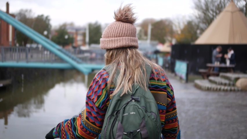 woman with colourful sweater, beanie hat and a backpack walking by te waterside playing with the fence in Bristol UK