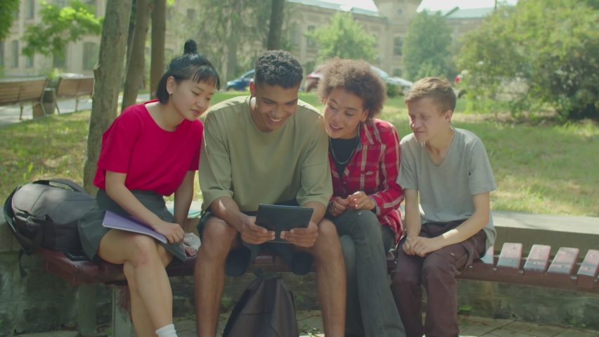 Positive beautiful diverse multicultural young people sharing digital tablet, networking and browsing social media content online while enjoying leisure, sitting on bench in public park.