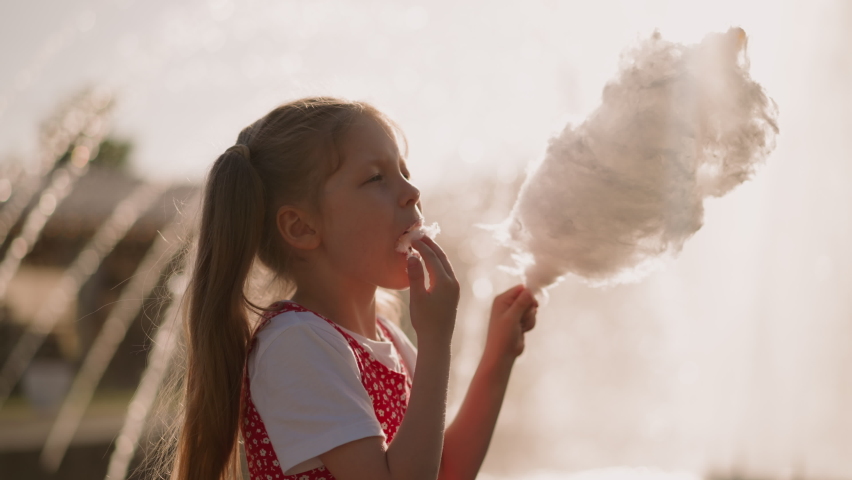Little girl puts piece of cotton candy into mouth and looks at sticky fingers against blurry fountain slow motion. Spending time in amusement park Royalty-Free Stock Footage #1092528157