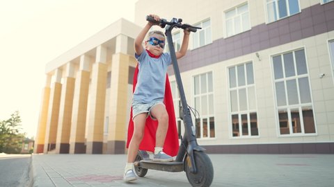 Child kid in superhero costume rides an electric scooter at school.Kid dream of travel and freedom.Schoolboy on an electric scooter in school yard.Child play in superhero costume and mask.Kid dream