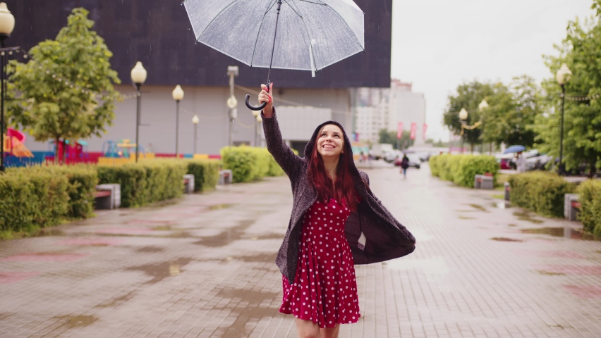 Young happy beautiful woman holding transparent umbrella high above head, standing in city park. Joyful pretty female enjoying rainy weather, wet climate on walk. Cheerful lady looking at downpour.