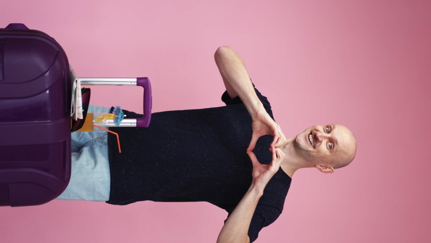 Holiday fun. Funny man. Love confession. Attractive excited guy with suitcase showing heart gesture on chest on pink background vertical.